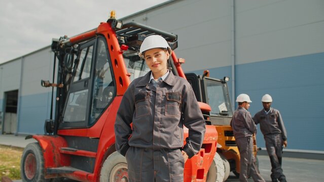 Beautiful girl wearing working uniform putting her hands in her pockets. In background two men of mixed race talking while standing near forklift. Workers waiting for goods to unload at factory.