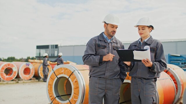 Busy Caucasian workers communicating while standing next to factory products. Pretty woman holding clipboard and speaking with her foreman. People wearing safety equipment during working at factory.
