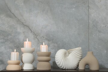 Candlesticks with burning candles and other decor on wooden table near light grey wall indoors,...
