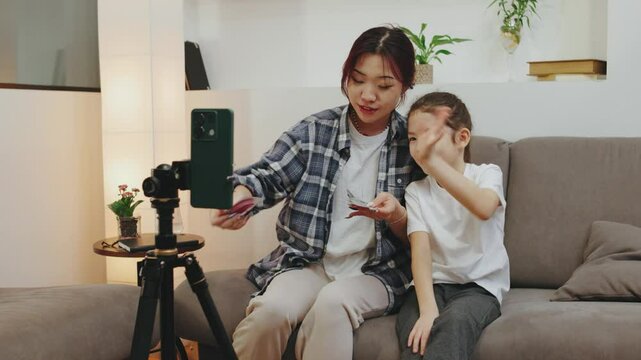 A mother and daughter in a cozy apartment record a video with a tripod. They wear under-eye patches, possibly creating social media content about makeup