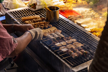 Street vendor grilling various meat skewers over open flames at a night market, hands shown turning the skewers with smoky atmosphere and glowing embers