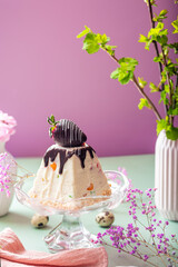 A luxurious cottage cheese Easter bread drizzled with chocolate and topped with a vibrant berry. Displayed on a glass stand with purple flowers and green leaves, creating a festive and elegant holiday