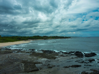 Dramatic storm clouds gather over rugged coastline where waves crash against dark volcanic rocks and sandy cove lined with lush vegetation, showcasing wild, moody beauty of Costa Rica's Pacific shore
