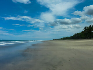Vast, wet sandy shore stretches alongside ocean, reflecting partly cloudy blue sky, as gentle waves roll in towards palm-fringed tropical coastline, capturing a serene, empty beach scene in Costa Rica