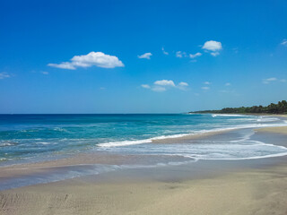 Fototapeta premium Gentle turquoise ocean waves roll onto pristine sandy beach under vibrant blue sky dotted with fluffy clouds. Scenic Costa Rican coastline stretches into distance, bordered by lush tropical vegetation