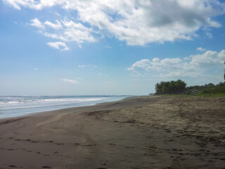 Wide, dark sand beach, marked with footprints, extends along tropical Costa Rica coastline. Gentle ocean waves lap the shore under bright blue sky dotted with fluffy white clouds and lush vegetation