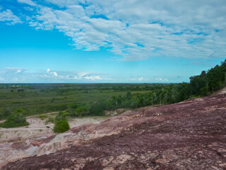 From reddish eroded cliffs, a vast landscape of green vegetation unfolds, stretching towards distant blue ocean horizon under a partly cloudy sky within the Pataxo Indigenous Reserve, Bahia, Brazil