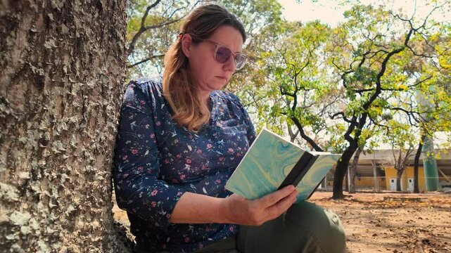 Adult caucasian woman with Photochromic Bifocal Reading Glasses sits under tree in a park and read a book. Relax in Nature