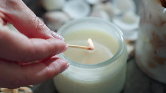 Close up of woman with polished nails lighting white candle with matchstick, flame touching candle wick, surrounded by seashells and starfish on marble table