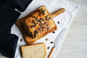 Loaf of bread with pumpkin seeds close-up on wooden board and cotton towel near knife on grey background
