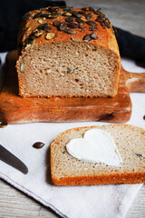 Slice of bread with butter heart next to loaf of bread with pumpkin seeds on wooden board and knife on grey background