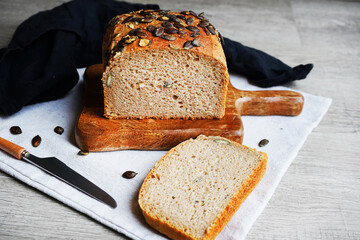 Loaf of bread with pumpkin seeds close-up on wooden board and cotton towel near knife on grey background