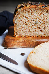 Loaf of bread with pumpkin seeds close-up on wooden board and cotton towel near knife on grey background