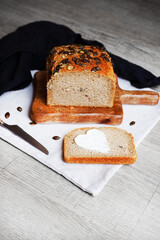 Slice of bread with butter heart next to loaf of bread with pumpkin seeds on wooden board and knife on grey background
