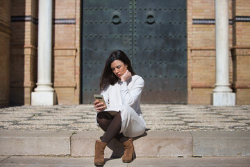Young and beautiful Latin brunette woman is sitting on a staircase looking at her mobile phone. The...