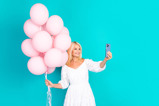 Smiling woman holding pink balloons while taking a selfie against a teal background in a white stylish dress