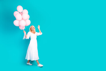 Elegant mature woman holding pink balloons while smiling in a white dress on a vibrant teal background