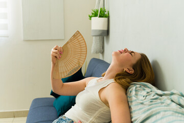 Stressed woman sitting on sofa at home using hand fan during summer heat wave