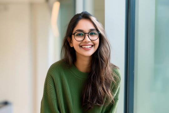 A close up head shot portrait of a preppy, young, beautiful, confident and attractive Indian Asian woman in a green sweater and spectacles in a classroom or office. She is smiling happily