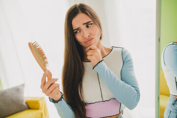 Young woman examining her hairbrush indoors at home, expressing curiosity or concern in a casual...