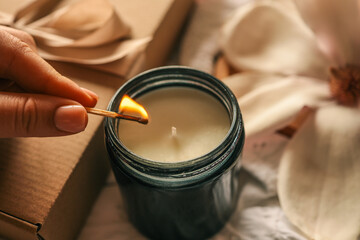A woman lights a soy candle in a dark glass jar with a matchstick, surrounded by a kraft gift box, blooming magnolia flowers, and a cup of coffee on soft embroidered fabric in warm light