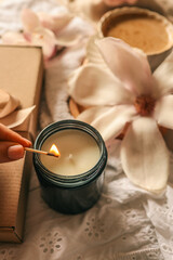 A woman lights a soy candle in a dark glass jar with a matchstick, surrounded by a kraft gift box, blooming magnolia flowers, and a cup of coffee on soft embroidered fabric in warm light