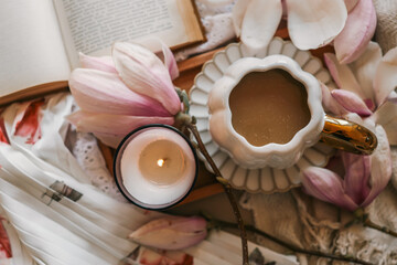 Top view of a cozy scene with a lit candle, scalloped coffee cup, magnolia flowers, and an open book on soft fabric in warm tones