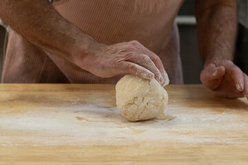 An unrecognizable Caucasian adult male rolling a ball of raw pastry dough on a floured wooden kitchen countertop.