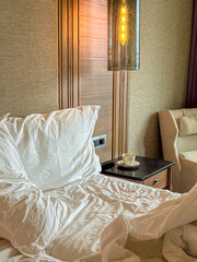 Unmade bed with white linens, a glowing bedside lamp, and a coffee cup on a nightstand. Ideal for travel ads, lifestyle blogs, and hotel interior concepts.