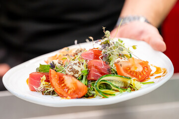 A chef presents a beautifully arranged plate of fresh salad featuring sliced tomatoes, tuna, mixed greens, and drizzled dressing. The vibrant colors and elegant presentation highlight a gourmet dish.
