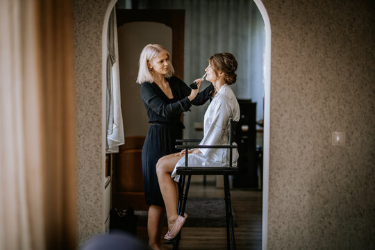 Makeup artist applying makeup to bride sitting on chair in cozy room during wedding morning preparation, natural light and calm atmosphere..