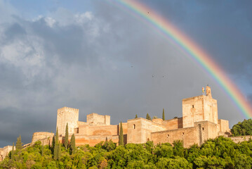 Alhambra castle under rainbow, birds flying in cloudy sky, Granada, Spain