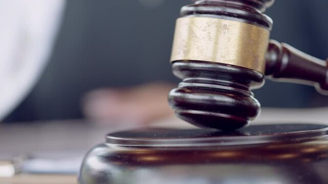 A judge is using a wooden gavel with a gold band to make a decision. The gavel is placed on a wooden surface, possibly a desk or a podium. The judge is likely presiding over a court case or hearing