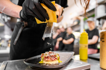 A chef in black gloves grates cheese over a meticulously plated dish, showcasing a refined culinary...
