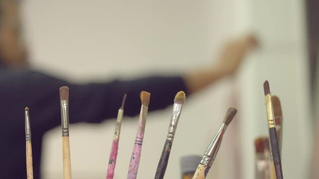 A man is standing behind a table with many paint brushes on it. The brushes are of different sizes and colors