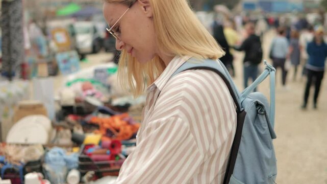 Young blond woman browsing vintage market stalls, wearing striped shirt and blue backpack, rummaging through secondhand items with playful curiosity and bright smile.Europe travel.