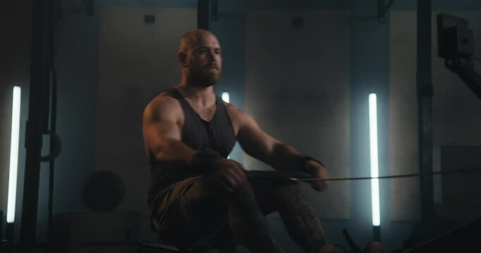 Bearded muscular man in a tank top working out on a rowing machine in a modern gym