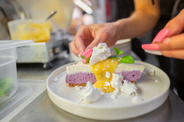 A professional chef decorates a layered dessert with fresh mint leaves and crumbled cheese. The dessert features purple and white mousse topped with yellow sauce, on a clean plate.