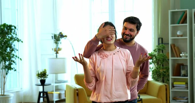 Indian Asian young handsome husband surprises his beautiful wife by closing her eyes and giving a gift box while standing in a stylish modern living room, wife smiles expressing joy and excitement