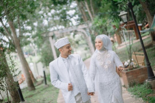 Happy Muslim wedding couple walking and posing in park, wearing traditional white attire. Outdoor Islamic marriage photography, natural and romantic prewedding style.