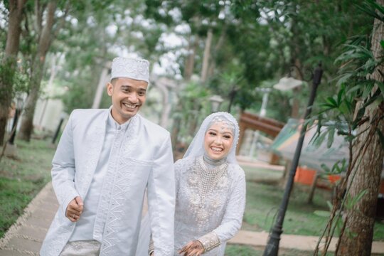 Happy Muslim wedding couple walking and posing in park, wearing traditional white attire. Outdoor Islamic marriage photography, natural and romantic prewedding style.