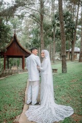 Fototapeta premium Romantic Muslim wedding couple in white standing in a green garden. Peaceful and elegant outdoor moment, perfect for Islamic, cultural, and love-themed wedding content
