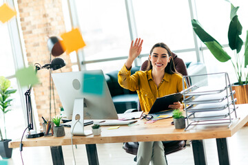 Young professional businesswoman in a modern office workspace waving hello, showcasing a friendly...