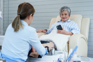 Obraz premium Asian female spa worker performing foot treatment for senior female customer relaxing on massage chair holding smartphone while smiling during wellness session inside cozy modern beauty salon