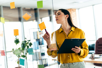 Young businesswoman in bright office analyzing ideas on sticky notes while holding a clipboard,...