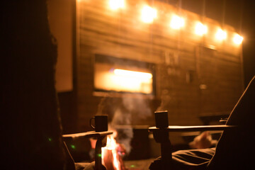 Сozy outdoor scene at night with two steaming mugs placed on wooden tables near glowing fire. The warm glow of string lights illuminates a wooden cabin in the background, creating a peaceful ambiance.