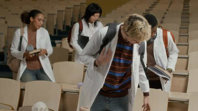 Rack focus shot of multiethnic group of young medical students walking into lecture hall then sitting down at desk in rows while professor marking attendance at beginning of college lesson