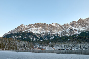 Obraz premium Twilight Alpine Wonderland: Eibsee Lake Reflects Snow-Capped Zugspitze Mountain Amidst Illuminated Pine Forest Enchanting Winter Landscape Captures Serene Beauty of German Ski Resort at Dusk.