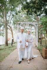 Muslim couple wearing elegant white wedding attire, walking together in a lush green garden. Perfect for prewedding, love, romance, and marriage celebration concepts.
