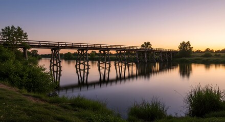 Fototapeta premium Wooden bridge reflecting beautifully on the calm river during a vibrant sunset, creating a peaceful and serene atmosphere amidst the surrounding natural landscape